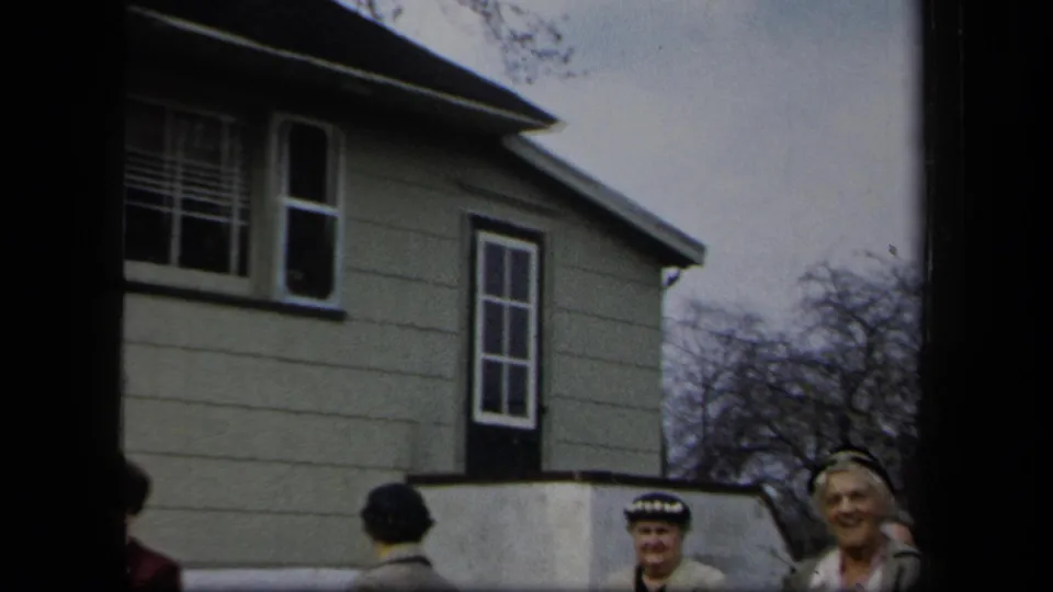 Two adult women stand near a residential-looking house exterior in New York, with a low wall, window, and bare trees in view.