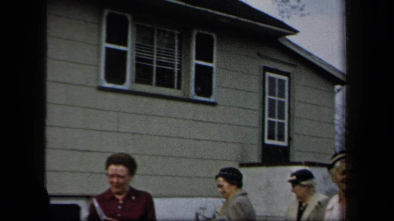 Women in period clothing walk past a light-sided residential house in 1958 New York home-movie footage.