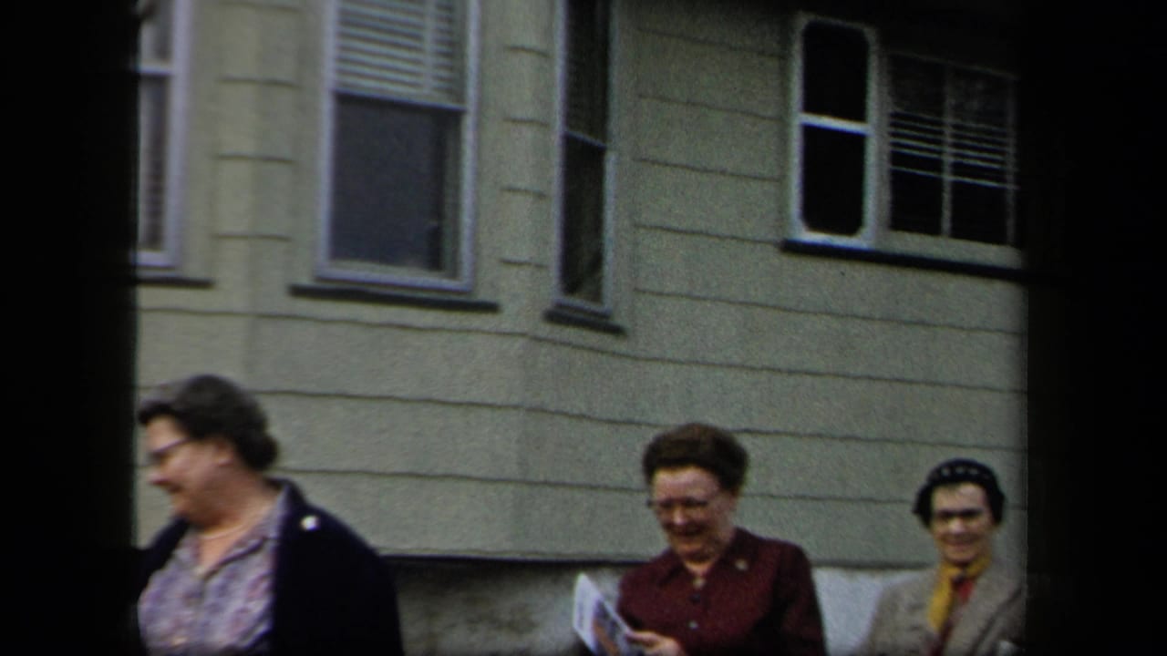 A group of women in coats and hats moves past a residential facade, with handheld 8mm framing and soft focus.