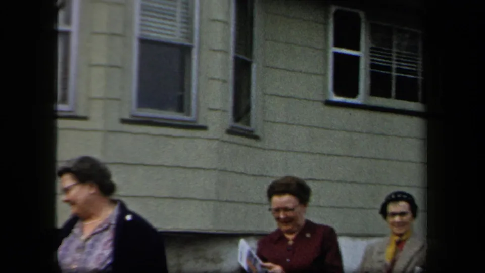 A group of women in coats and hats moves past a residential facade, with handheld 8mm framing and soft focus.