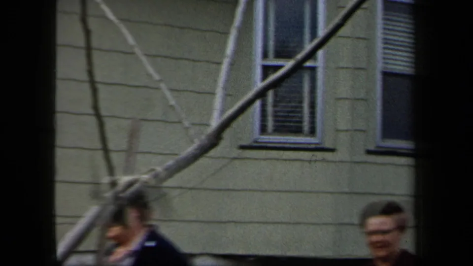 Soft 8mm frame of women passing a pale residential building, with bare branches visible in front.
