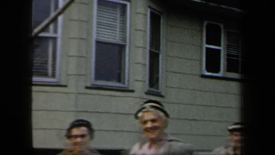 Women in 1950s-style hats and jackets walk beneath white-trimmed windows on a residential exterior.
