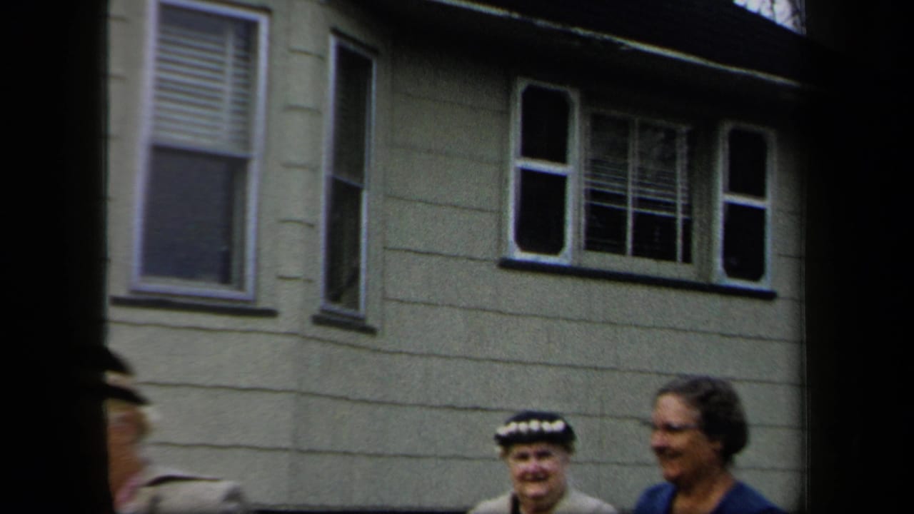 Women in period dress stand and smile outside a light clapboard house in 1958 New York home-movie footage.
