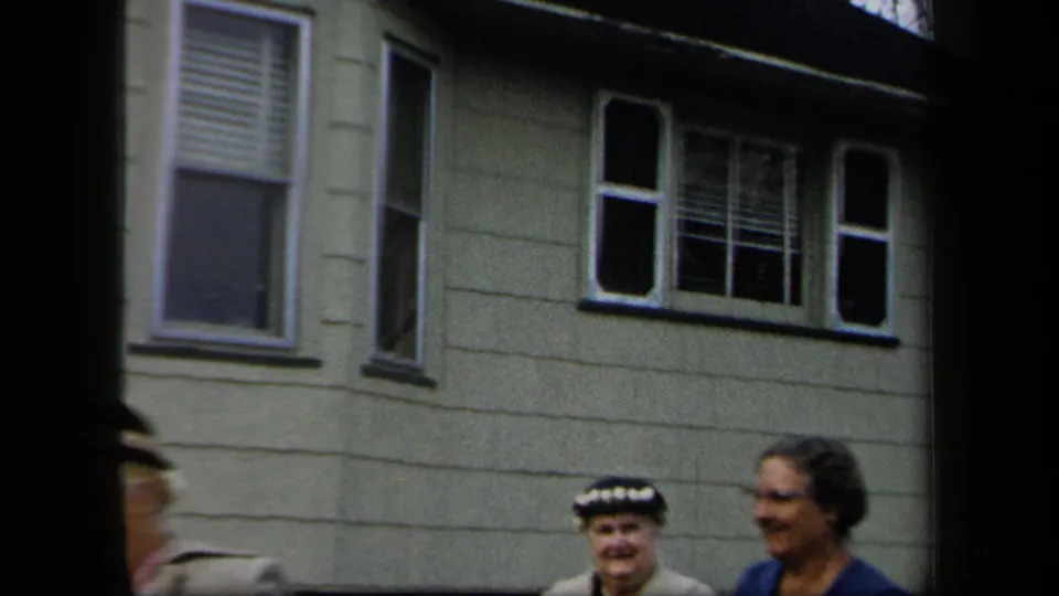 Women in period dress stand and smile outside a light clapboard house in 1958 New York home-movie footage.