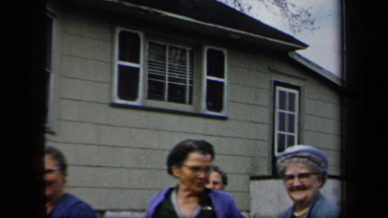 Women pass in front of a house exterior with shuttered windows.