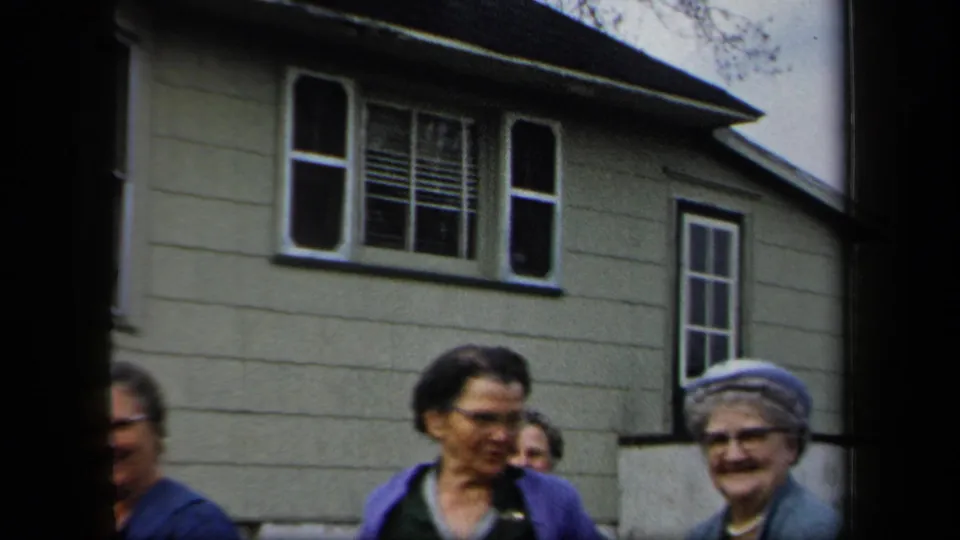 Several women move past the camera outside a light-colored house with shuttered windows in 1958 New York footage.