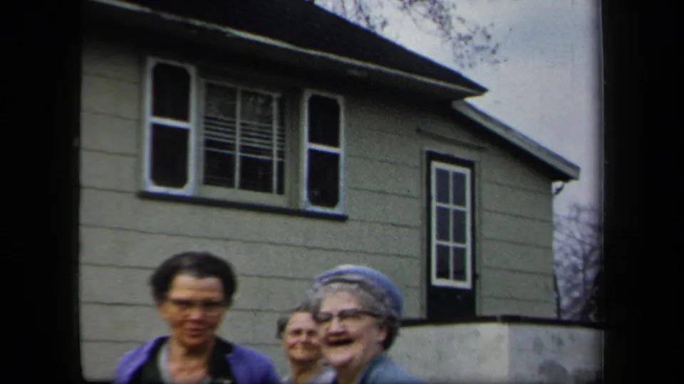 Three women stand close to the camera outside a clapboard house, with one wearing a blue hat in 1958 New York home-movie footage.