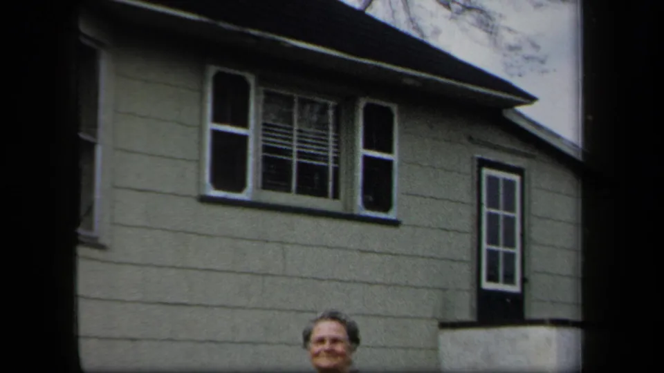 A woman is partly framed at the bottom of the shot below shuttered windows on a light clapboard house in 1958 New York footage.