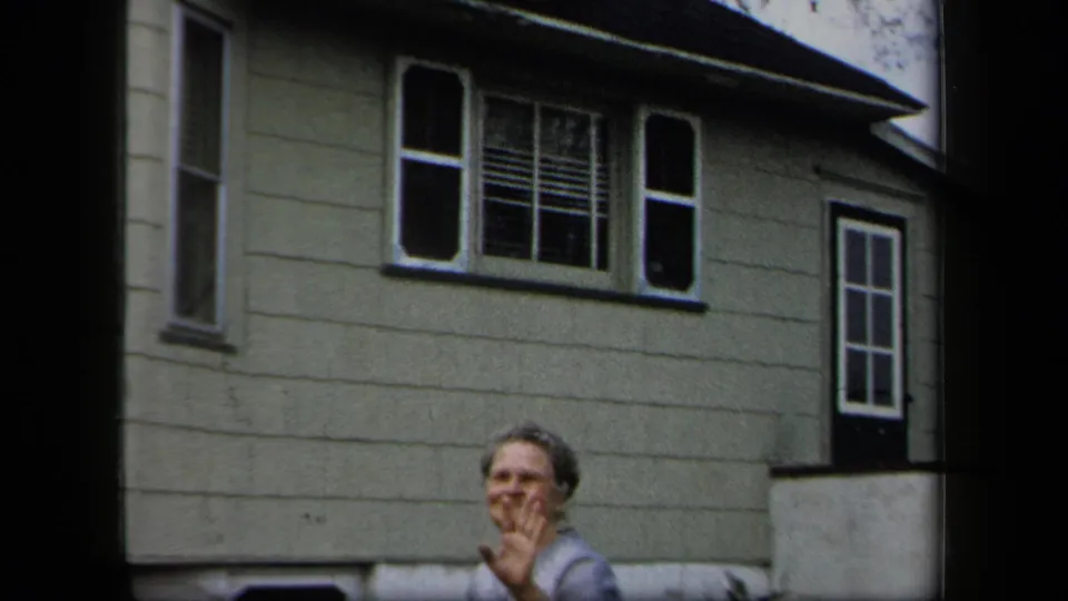 Adult woman smiles toward the camera outside a light-sided house in metadata-supported 1958 New York home-movie footage.