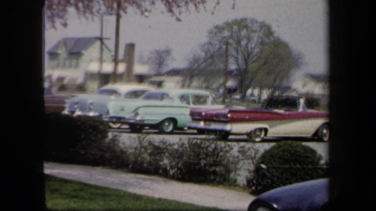 Classic cars are parked along a street with houses and trees in the background.