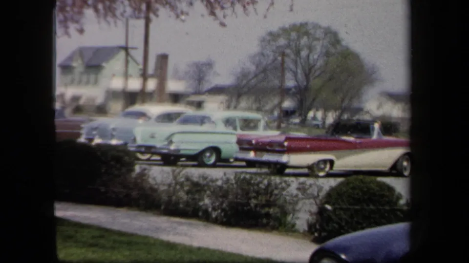 1958 New York frame with a red-and-white convertible, pale green sedan, other parked cars, low houses, and trees along a suburban street.