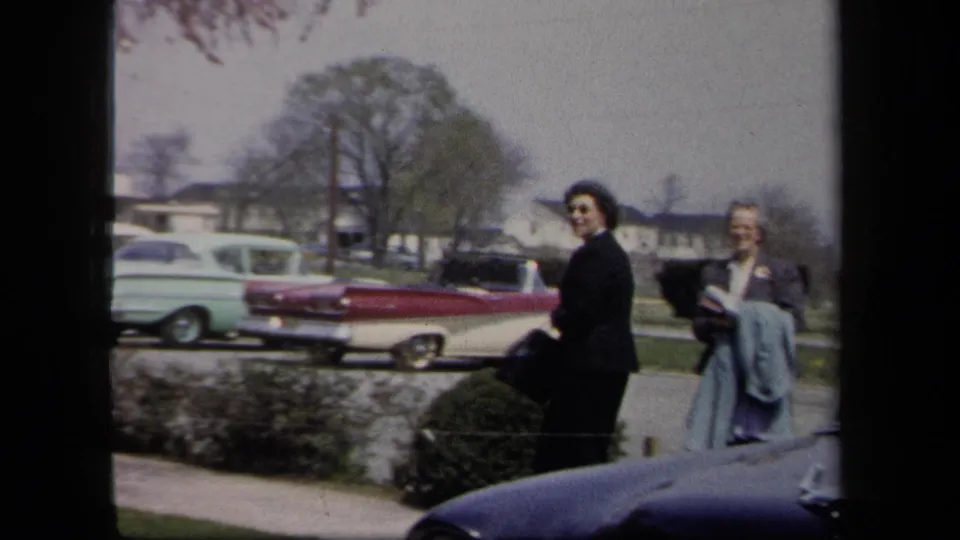 Two adult women in 1958 New York stand near shrubs and a sidewalk, with a red-and-white convertible and other parked cars behind them.