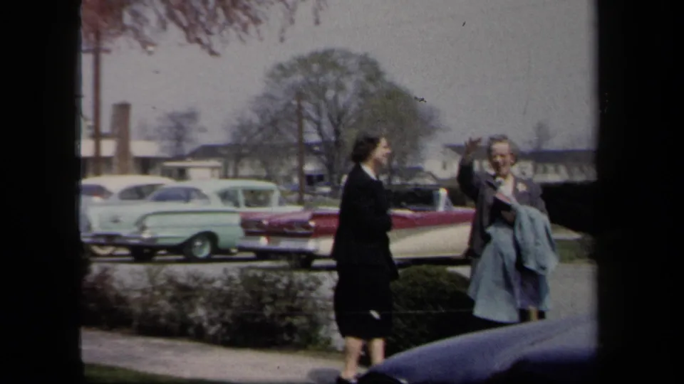 1958 New York home-movie frame of two adult women beside a sidewalk, one waving or gesturing, with mid-century cars and houses behind them.