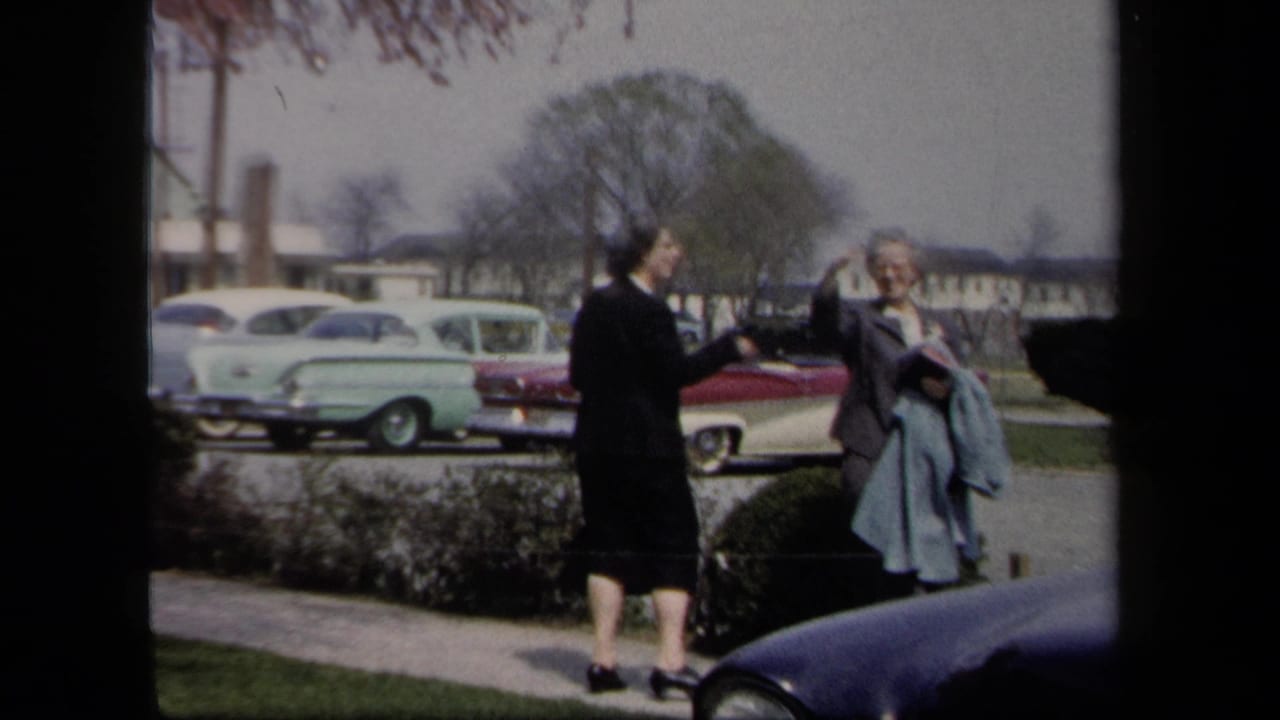 1958 New York home-movie frame showing two women in coats beside a parking lot with period cars and low buildings behind them.