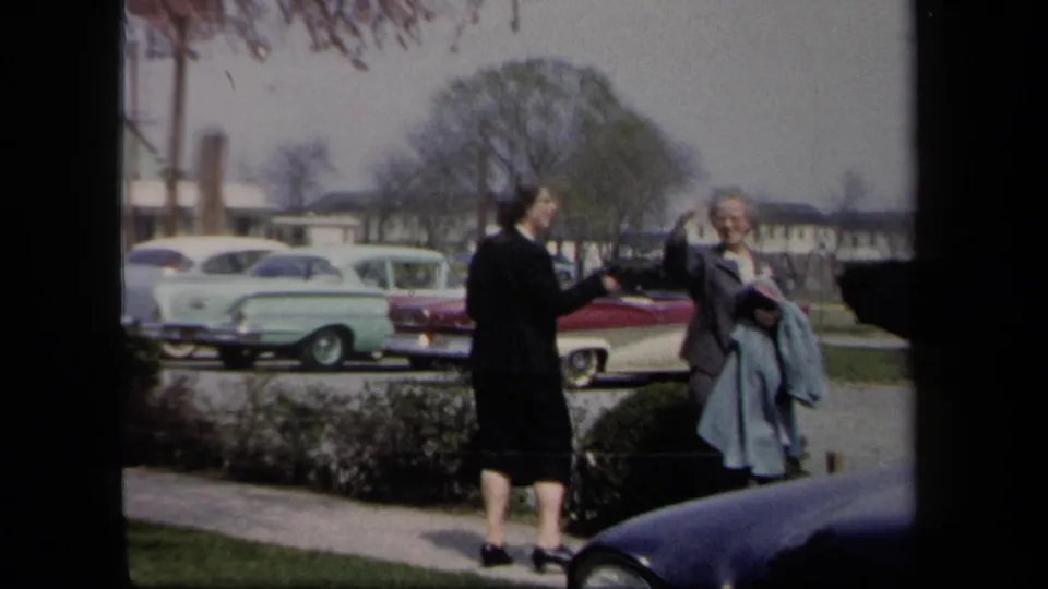 1958 New York home-movie frame showing two women in coats beside a parking lot with period cars and low buildings behind them.