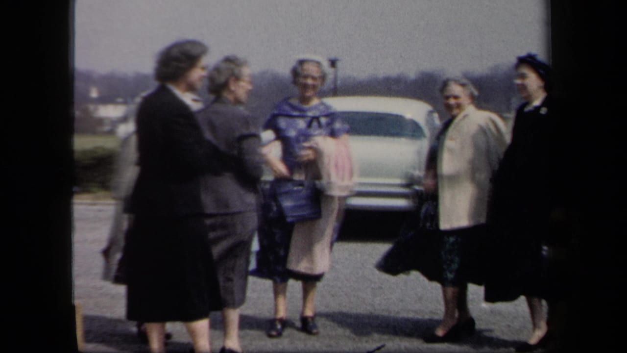 Six women in 1950s attire stand together near a pale car in a New York parking lot on a windy day.