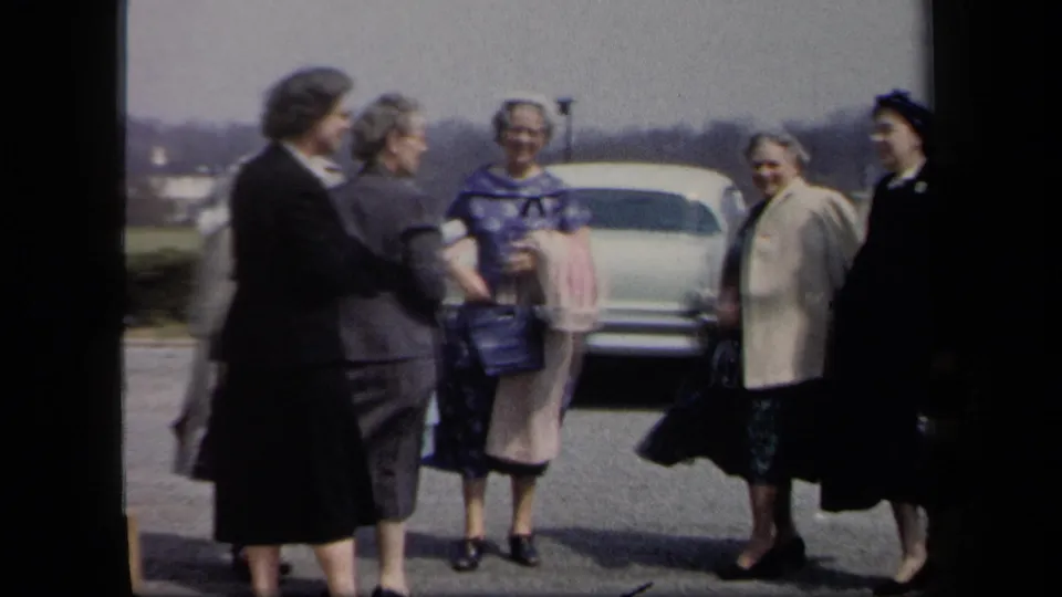 Six women in 1950s attire stand together near a pale car in a New York parking lot on a windy day.