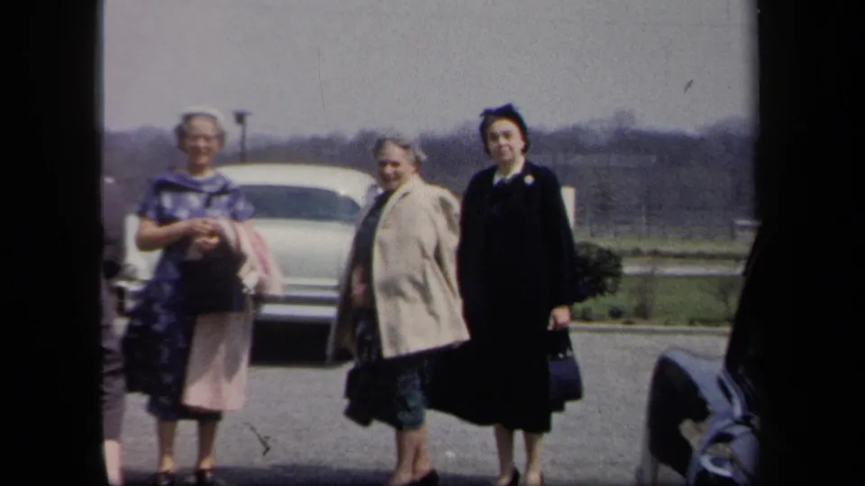 Three women in mid-century coats and dresses face the camera in a 1958 New York parking-lot home movie frame.