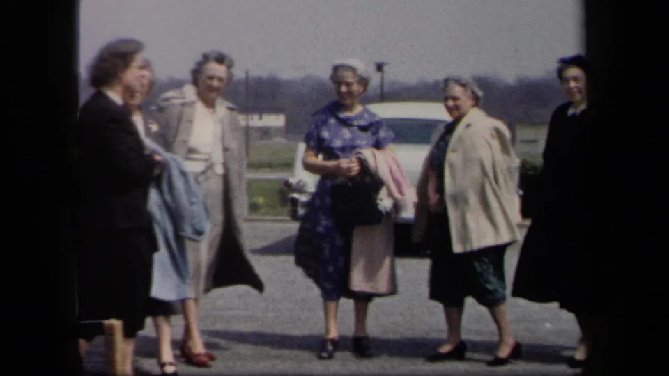 Six women in 1950s clothing pose in a New York parking lot beside a pale car, with coats held against the wind.