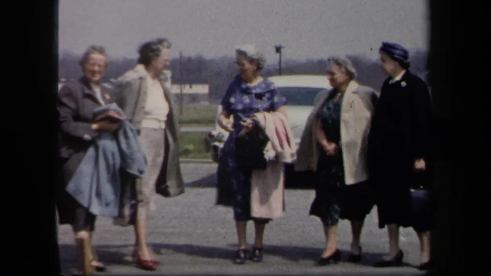 Five adult women in coats, dresses, hats, and handbags chat in a New York parking lot, consistent with 1958 metadata.
