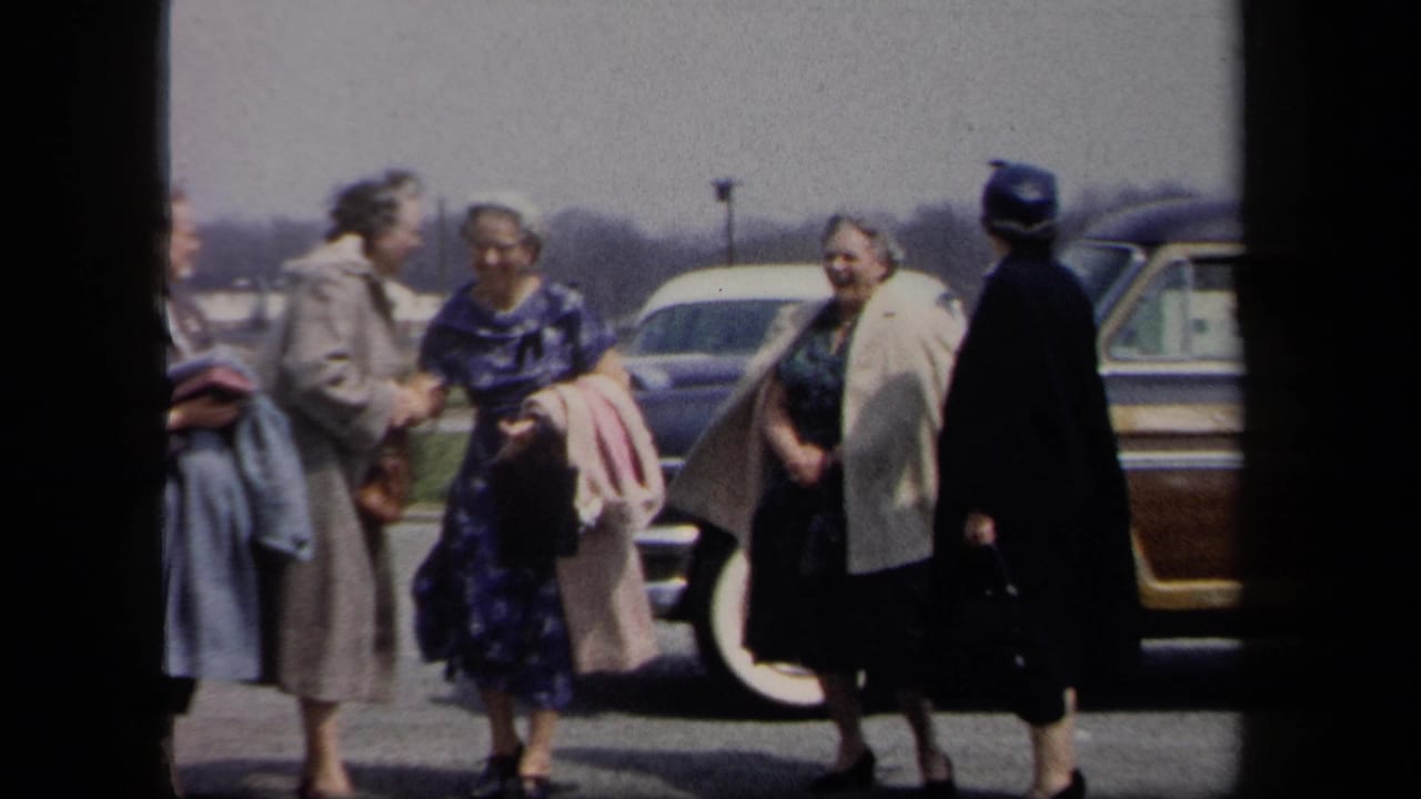 Women in mid-century coats and dresses stand together near a brown station wagon in a New York parking lot.
