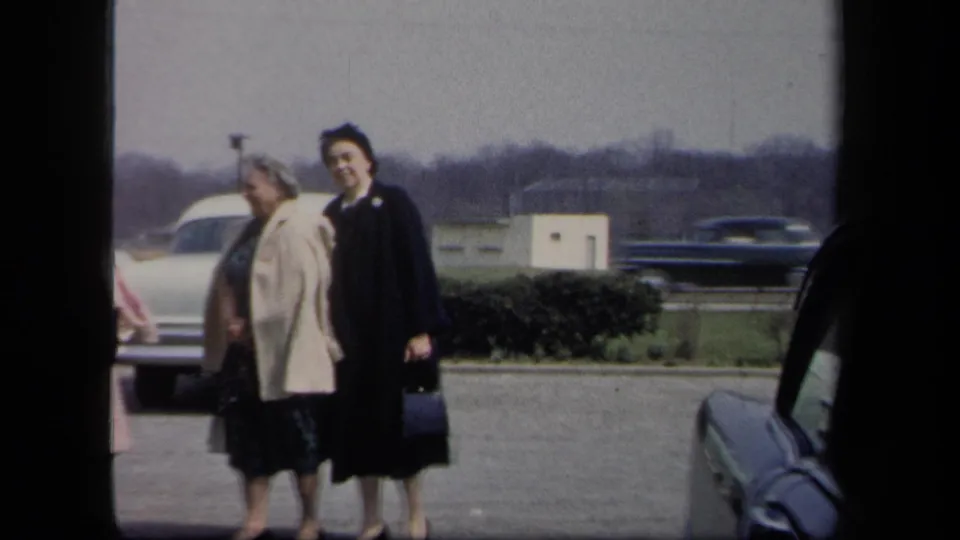 Two adult women, one in a light coat and one in a dark coat and hat, stand in a New York parking lot scene.