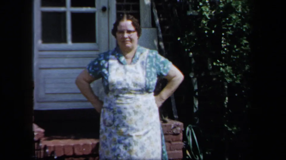1958 New York 8mm frame showing a woman in a floral dress and apron standing by a white door and brick garden edging.