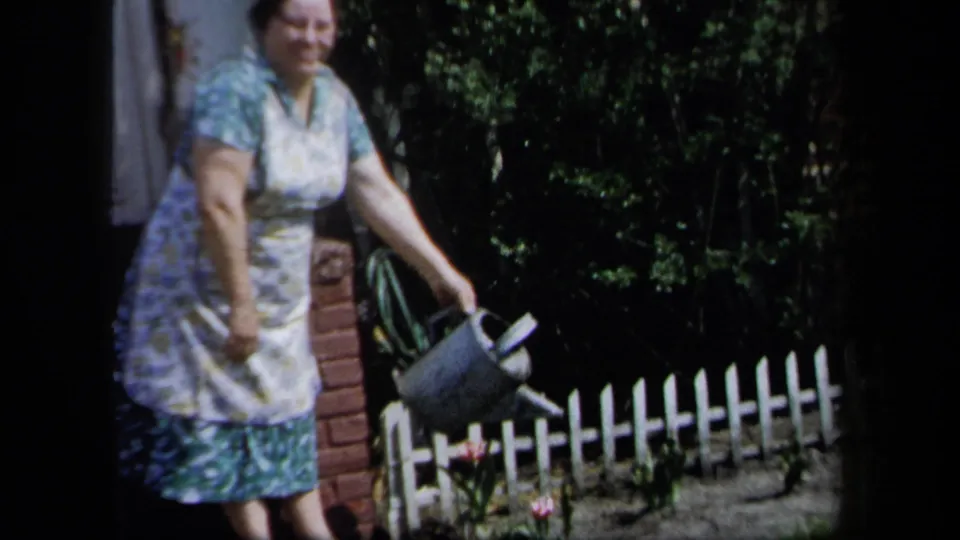 1958 New York 8mm frame showing a woman in a floral dress and apron watering flowers with a metal can near a white picket fence.