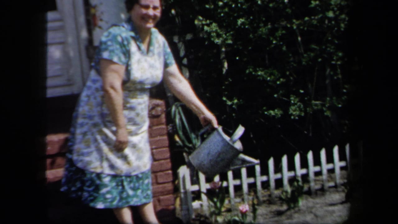 In 1958 New York home-movie footage, a woman in a floral dress and apron holds a metal watering can beside a garden fence.