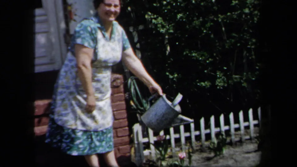 In 1958 New York home-movie footage, a woman in a floral dress and apron holds a metal watering can beside a garden fence.