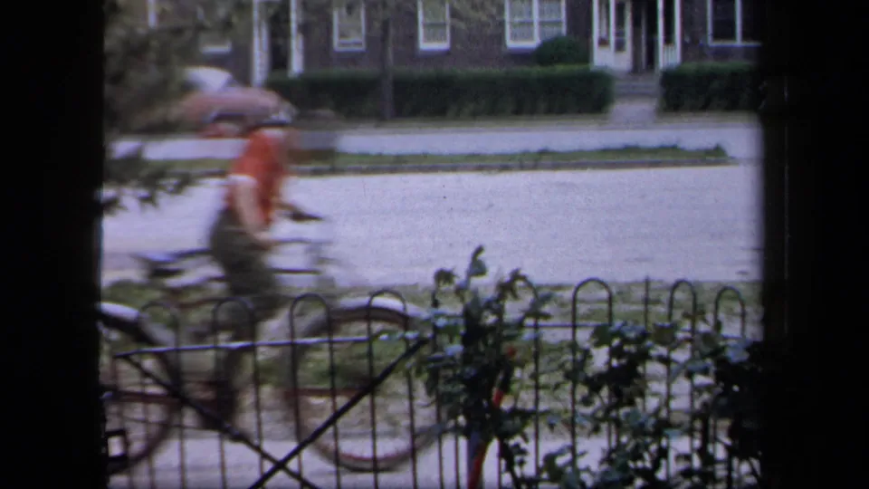 1958 New York 8mm frame showing a motion-blurred bicyclist beyond a low fence, with sidewalk, garden plants, street, and houses visible.