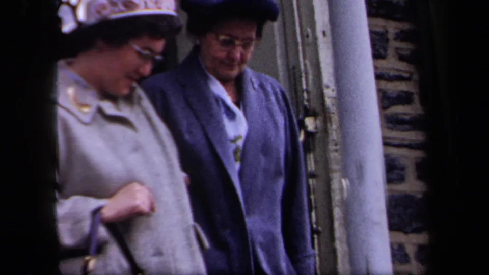 1958 New York home-movie frame showing two older women in mid-century coats and hats descending exterior steps beside a stone wall.