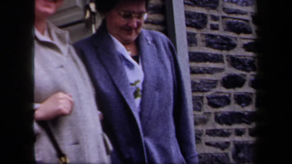 1958 New York home-movie frame of older women in dress coats near exterior steps, with one smiling beside a stone wall.