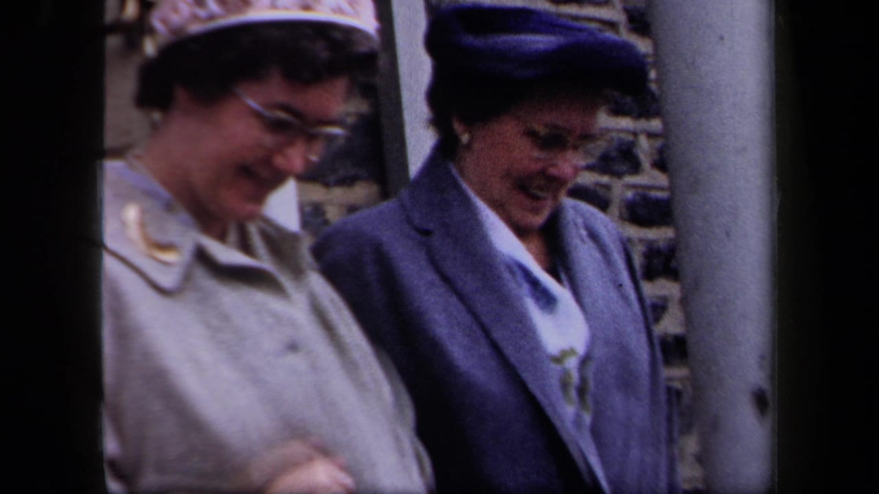 Two middle-aged women in 1950s New York walk close together outside a stone building, wearing hats and coats.