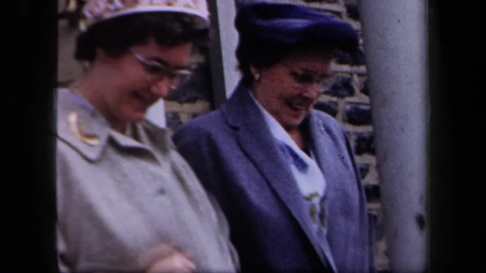 Two middle-aged women in 1950s New York walk close together outside a stone building, wearing hats and coats.