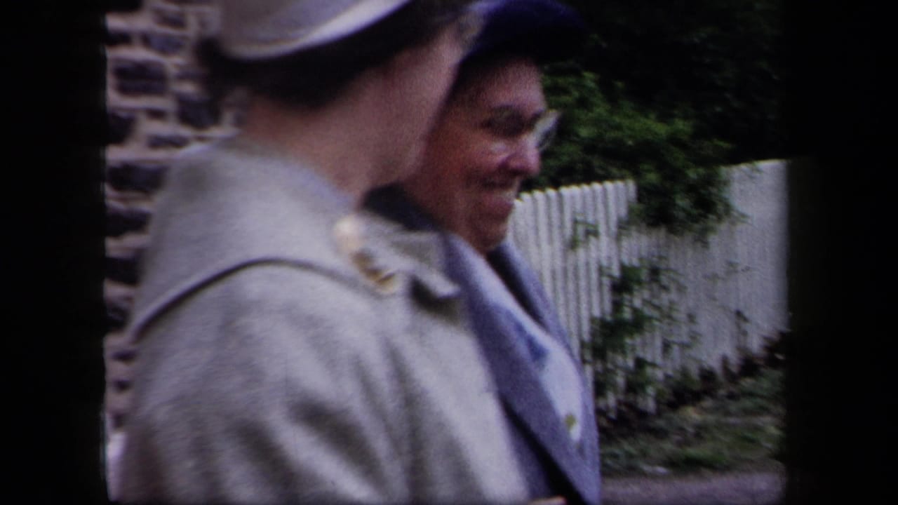 A woman in a light coat passes in front of another woman by a fence.