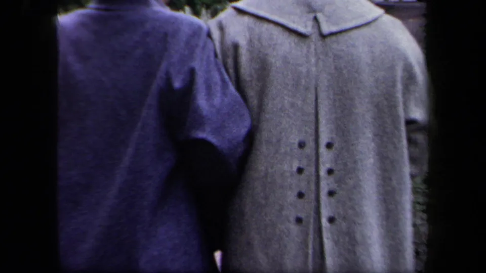 A close handheld frame shows the women’s mid-century coats and hats as they pass beside the stone building.