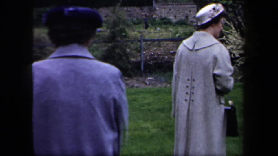 Two women in mid-century coats walk through a New York garden area, one wearing a patterned hat and carrying a handbag.