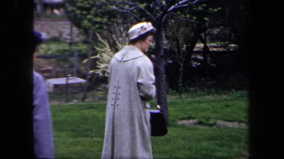 A woman in a light coat and patterned hat stands near the garden lawn in 1958 New York home-movie footage.