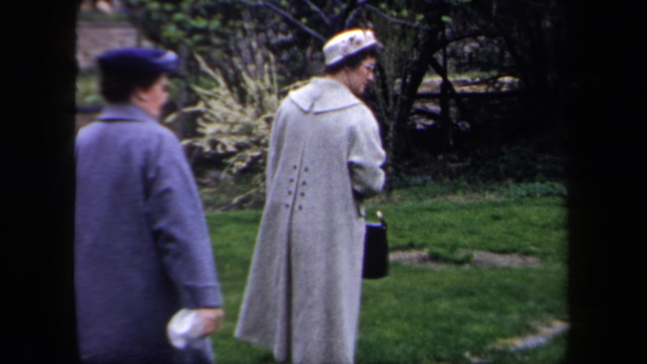 In a 1958 New York home-movie frame, two adult women in dress coats and hats walk through a green garden.