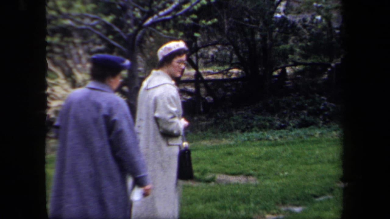 Two women pause on the lawn, one turning toward the camera with a handbag.