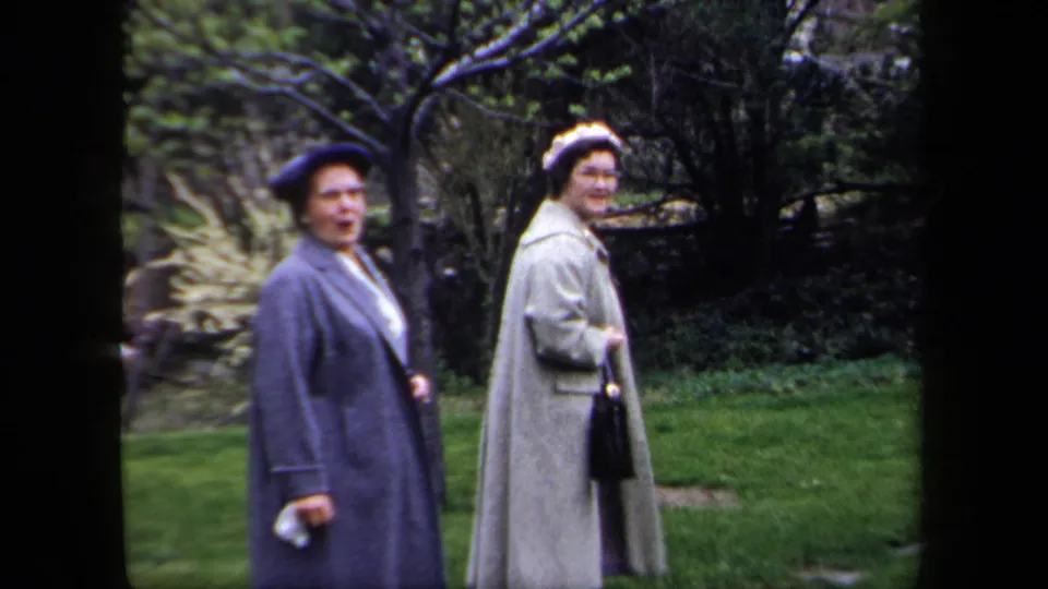 Two adult women in coats, hats, and gloves face the home-movie camera in a green garden setting.