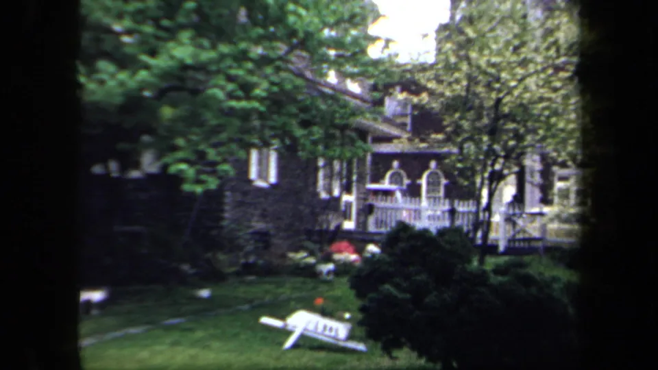 Home-movie view of a 1958 New York residential garden with a white-trimmed porch, trees, shrubs, lawn, and tipped chair.