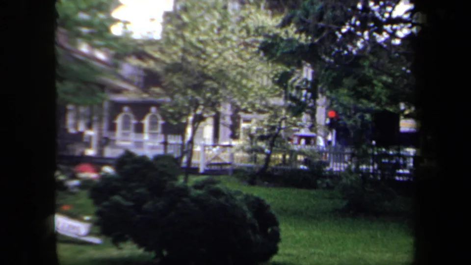 1958 New York residential yard frame with shaded grass, dense shrubs, tree branches, white porch trim, and a partially visible lawn chair.