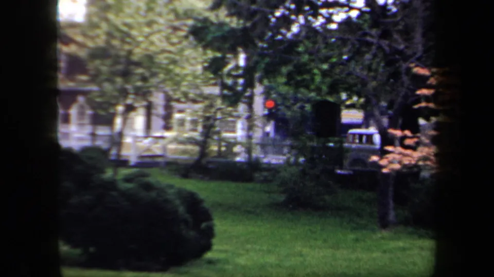 1958 New York home-movie frame looking through dark tree trunks across grass toward a fence, foliage, and a distant red traffic light.