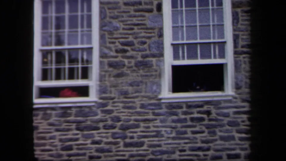 1958 New York home-movie view of a stone exterior with white sash windows, dark interiors, and a red object near one sill.