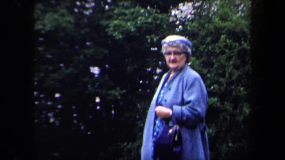 1958 New York home-movie frame of an older woman in a blue coat standing outdoors before dense greenery.