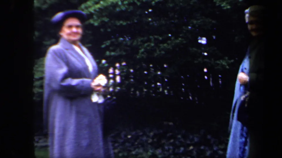 1958 New York home-movie frame showing older women in blue coats near greenery, with one woman wearing a hat and facing the camera.