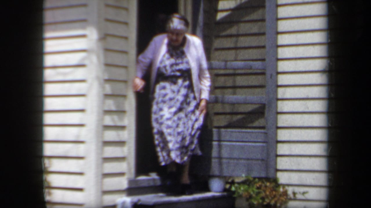 1958 New York home-movie frame showing an elderly woman in a floral dress and white cardigan leaving a yellow-sided house.
