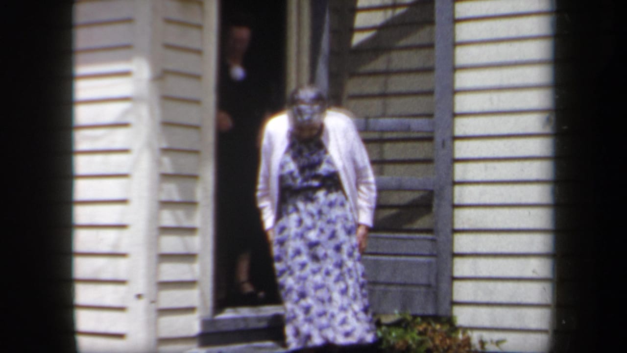 Woman in floral dress stands near the doorway of a yellow-sided house.